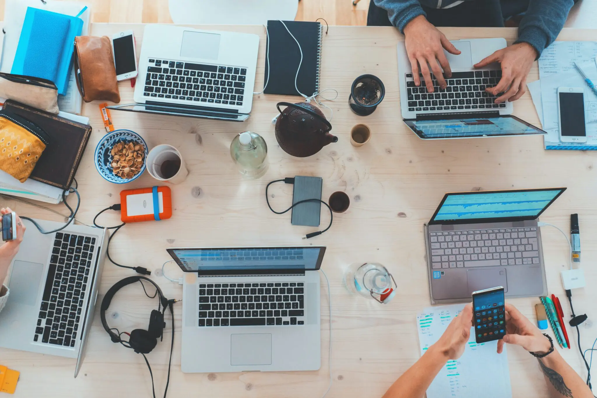 A desk covered in laptops, notebooks, headphones and a teapot