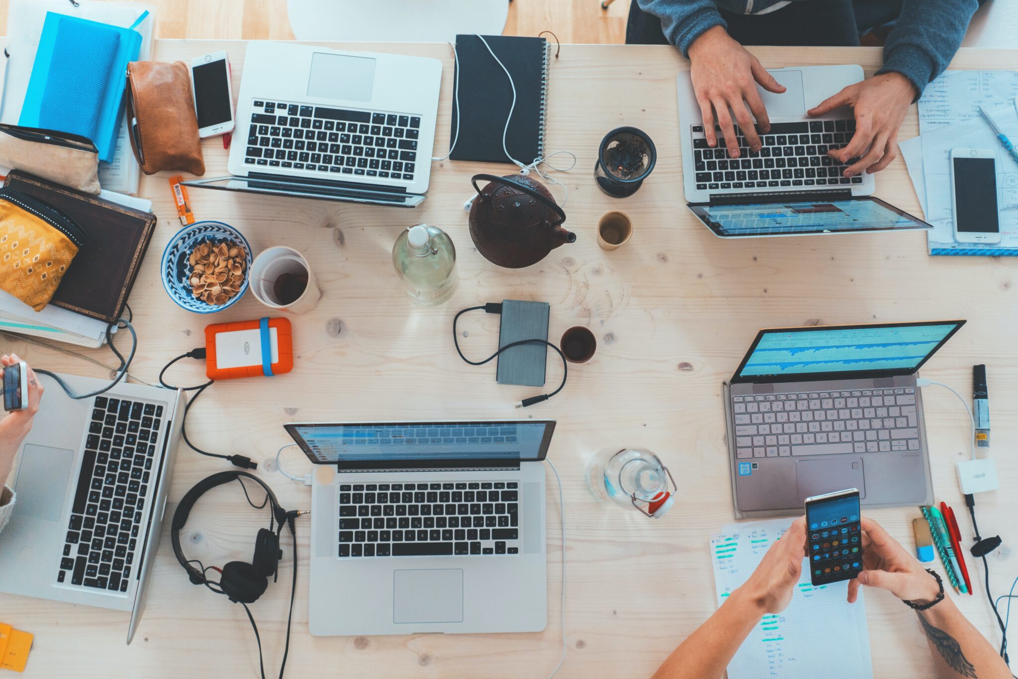 A desk covered in laptops, notebooks, headphones and a teapot